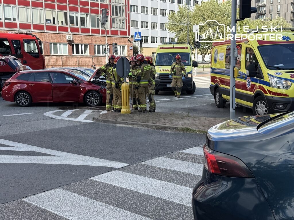 Czterech strażaków bada uszkodzony czerwony samochód osobowy na skrzyżowaniu, obok stoi ambulans.