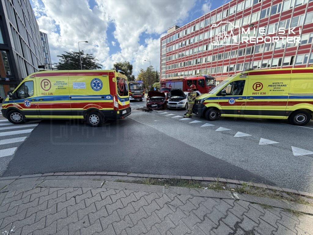 Dwa karetki pogotowia z włączonymi sygnałami zatrzymane na skrzyżowaniu obok uszkodzonego samochodu osobowego.