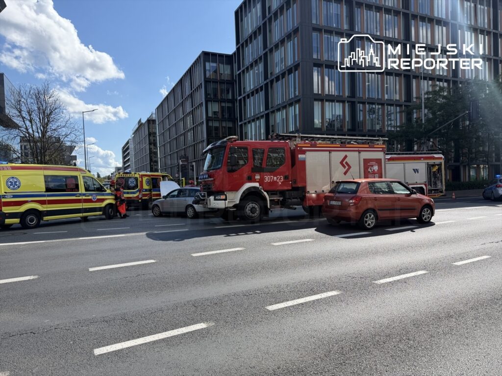Zespół ratunkowy w żółtych ambulansach oraz wóz strażacki blokują ruch na ulicy w otoczeniu nowoczesnych budynków biurowych.