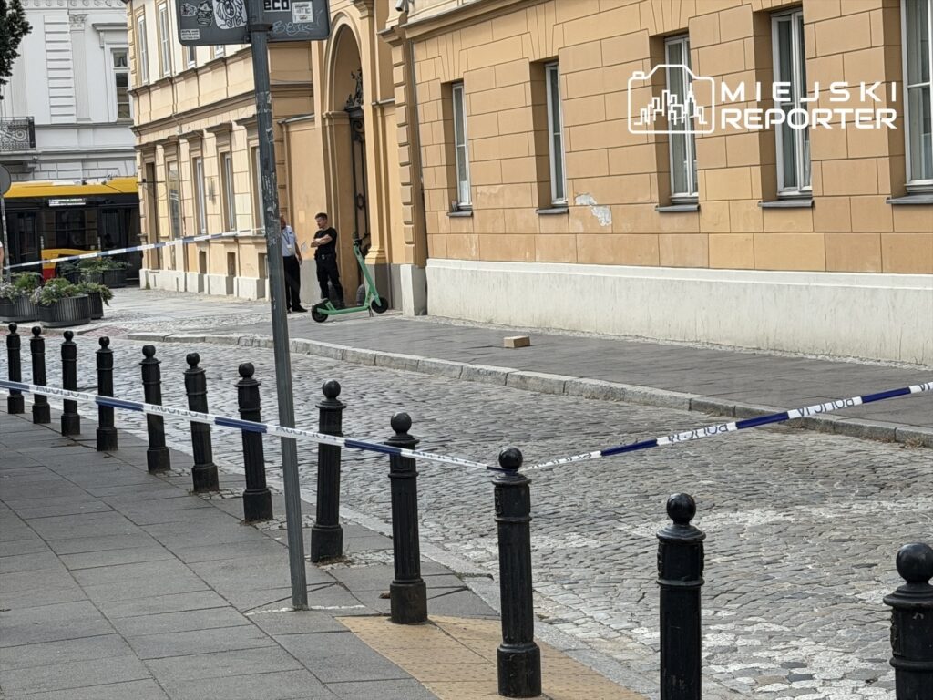 Na chodniku przy budynku dwóch policjantów rozmawia obok oznakowanego skuterka, a teren jest zabezpieczony taśmą.