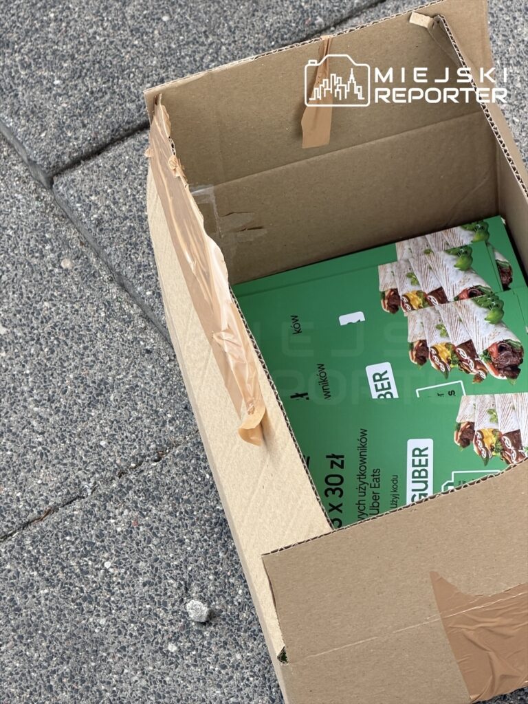 Otwarta tekturowa skrzynka leży na chodniku, zawierająca zielone opakowania z jedzeniem.