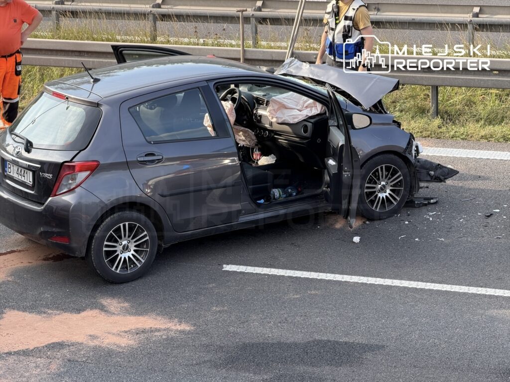 Czarny samochód osobowy z uszkodzoną tylną częścią stoi na pasie drogi, obok pracownicy służb ratunkowych.