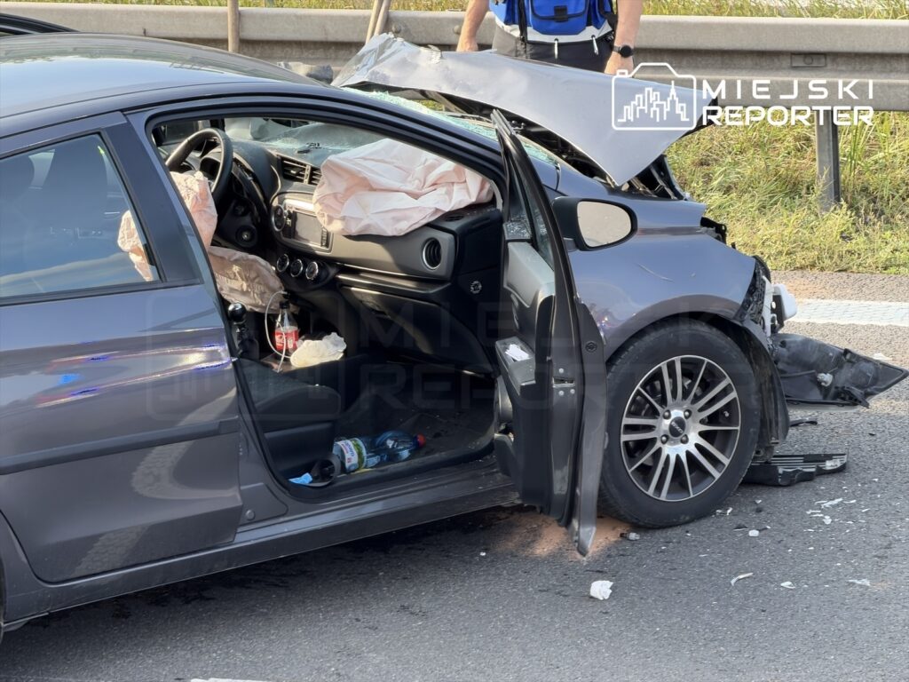 Uszkodzony samochód osobowy z otwartymi drzwiami, w środku poduszki powietrzne, na drodze wzdłuż barierki.