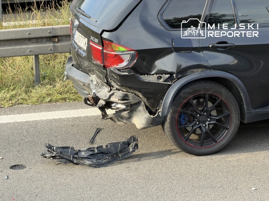 Czarny SUV z uszkodzeniami tyłu oraz odpadniętym zderzakiem stoi na pasie ruchu, obok widać trawę.