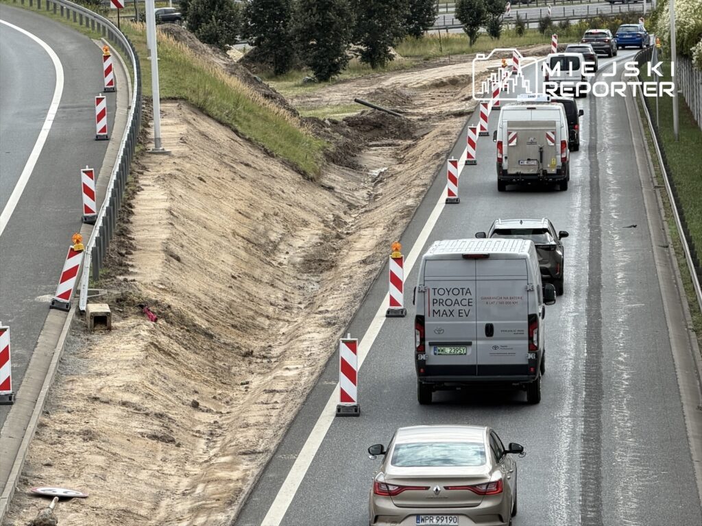 Ruch pojazdów na drodze krajowej, z oznaczeniami robót drogowych i wykopem po prawej stronie.
