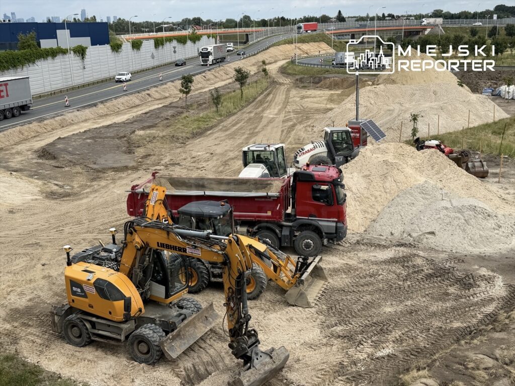 Na placu budowy znajdują się ciężkie maszyny, w tym żółty koparko-ładowarka i czerwona ciężarówka, otoczone piaskiem.