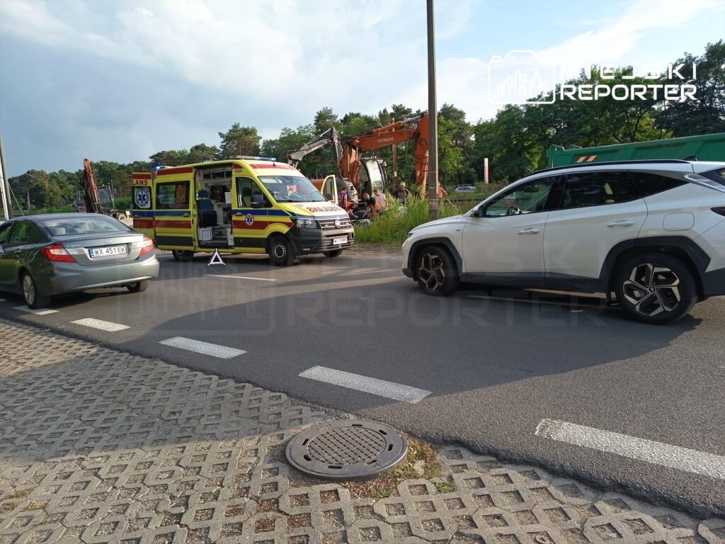 Ambulans z włączonymi sygnałami świetlnymi stoi na drodze obok dwóch samochodów osobowych, w tle widoczna koparka.