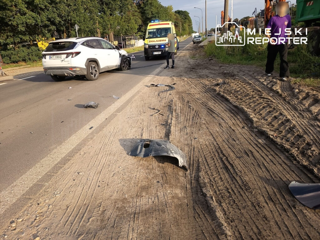 Na drodze leżą fragmenty karoserii, obok zatrzymany biały SUV oraz karetka pogotowia z personelem medycznym.