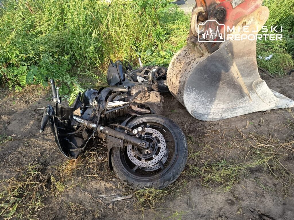Motocykl leży na ziemi obok koparki, z uszkodzoną konstrukcją i wygiętym kołem, otoczony wysoką trawą.