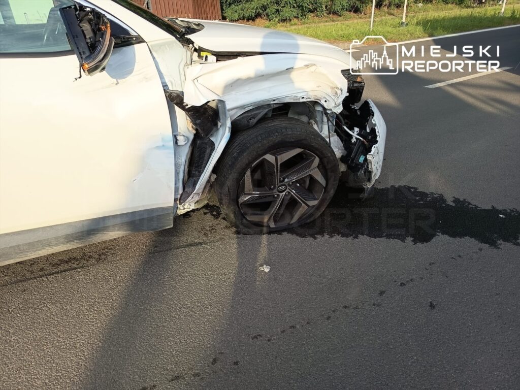 Uszkodzona strona białego samochodu osobowego z widocznymi zniszczeniami nadwozia i wyciekającym płynem na drodze.