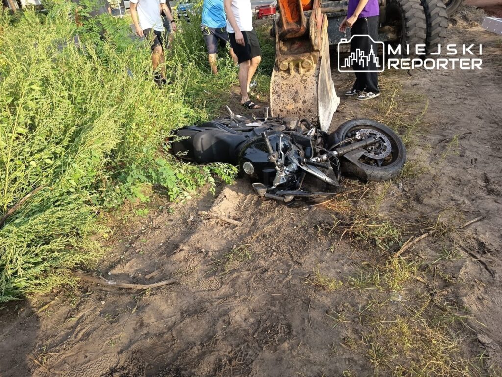 Zniszczony motocykl leży na ziemi wśród wysokiej trawy, obok koparki, a w tle stoją ludzie.