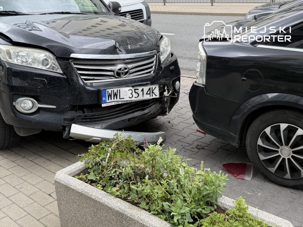 Czarny samochód osobowy z uszkodzoną przednią częścią stoi na chodniku obok innego pojazdu, w tle widać roślinność.