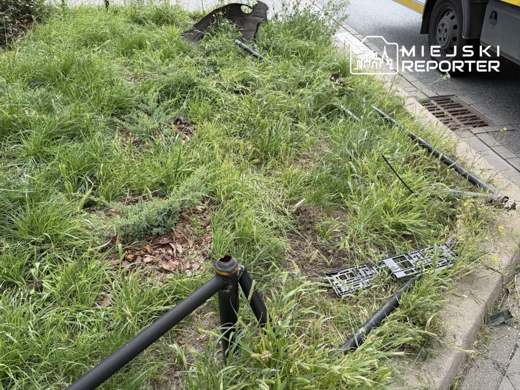 Zielona trawa i krzewy porastają teren, na którym leżą uszkodzone elementy ogrodzenia oraz fragmenty metalowych rur.