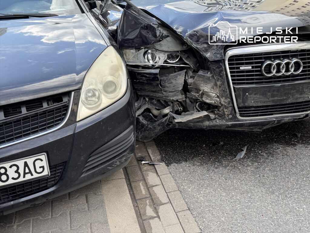 Czarne Audi z uszkodzoną przednią częścią zderza się z szarym samochodem osobowym na ulicy.