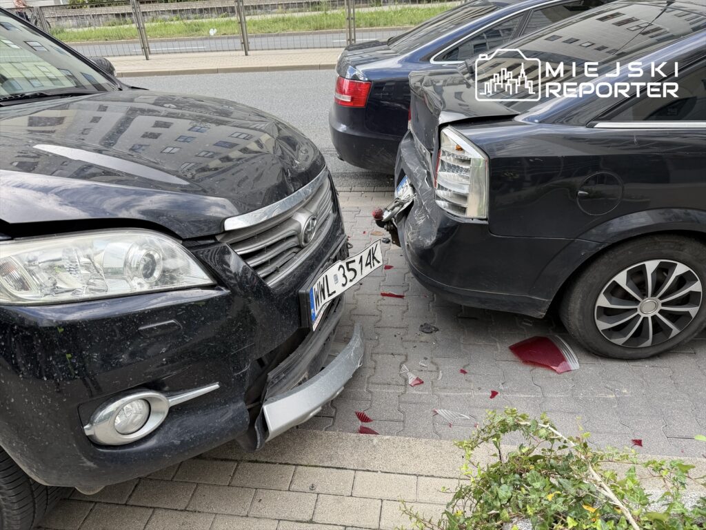Czarny samochód osobowy z uszkodzoną maską zderza się z innym pojazdem na zatłoczonej ulicy, w pobliżu chodnika.