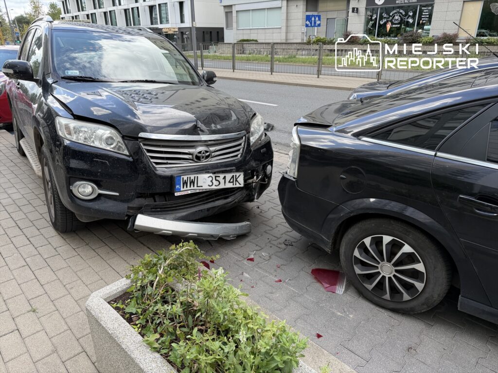 Czarny SUV stoi na chodniku z uszkodzoną przednią częścią, zderzając się z tyłem drugiego pojazdu w miejskim otoczeniu.