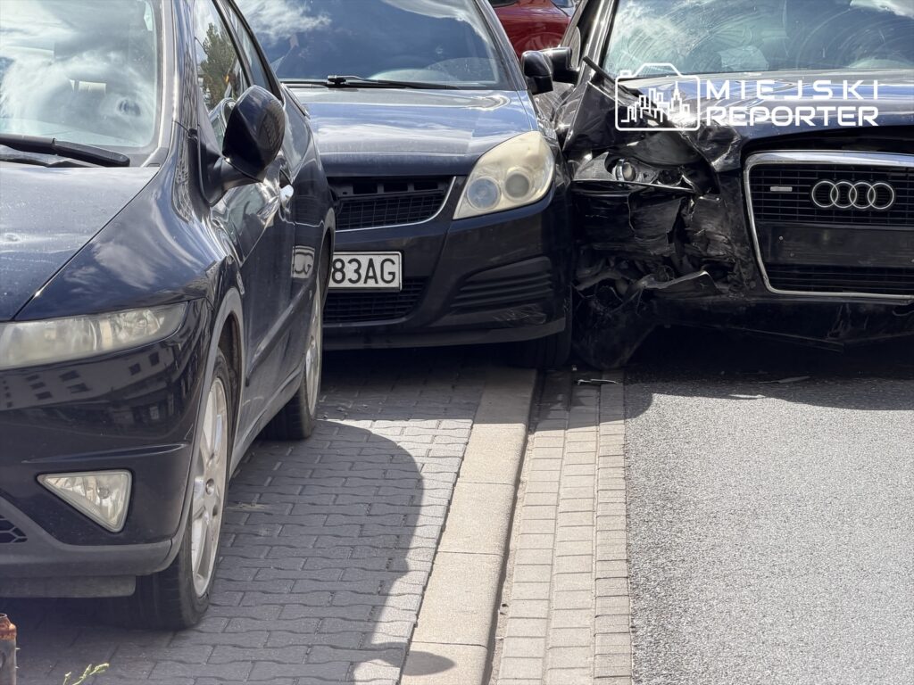 Czarny samochód Audi z uszkodzoną przednią częścią stoi na chodniku, zderzając się z innym pojazdem.