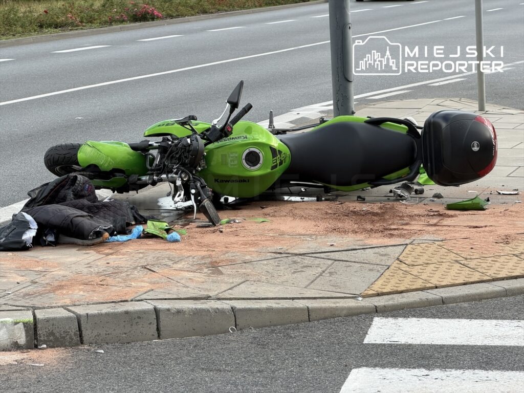 Zielony motocykl leży na boku na chodniku, wokół porozrzucane ubrania i przedmioty, w tle ruchliwa ulica.