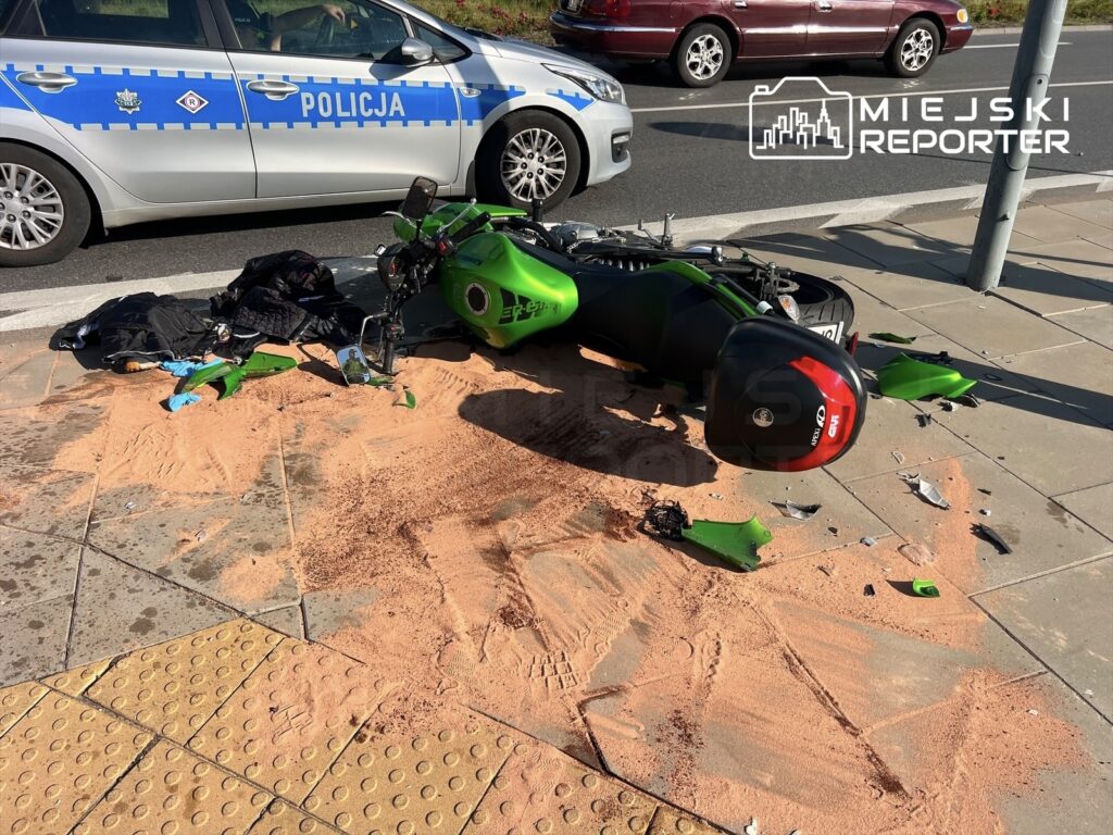 Zielony motocykl leży na chodniku obok rozrzuconych ubrań oraz piasku, a w tle widać policyjny radiowóz.
