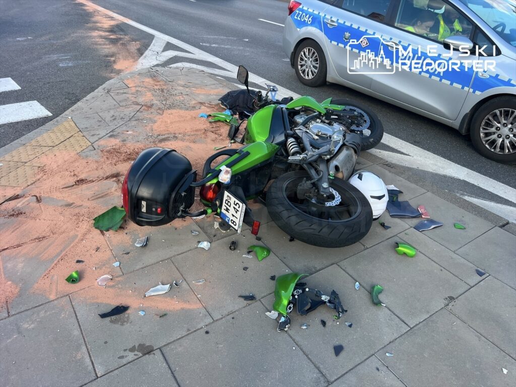 Motocykl w zielonym kolorze leży na boku, otoczony kawałkami szkła i innych odłamków, na chodniku przy ruchliwej ulicy.