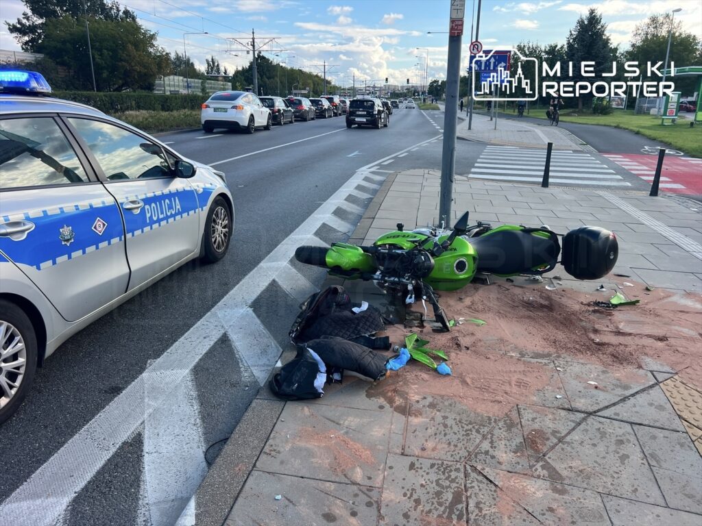 Zielony motocykl leży na boku na chodniku, obok rozrzucone ubrania i ślady na jezdni. Policja bada miejsce zdarzenia.