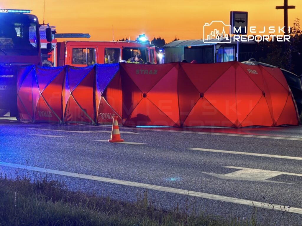 Czerwony namiot strażacki rozstawiony na drodze, obok pojazdów ratunkowych, w tle zachodzące słońce.