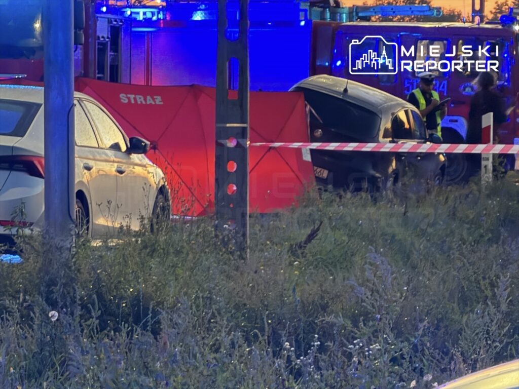 Na miejscu zdarzenia ratownicy pracują przy zderzonym samochodzie, otoczonym taśmą zabezpieczającą i namiotem.