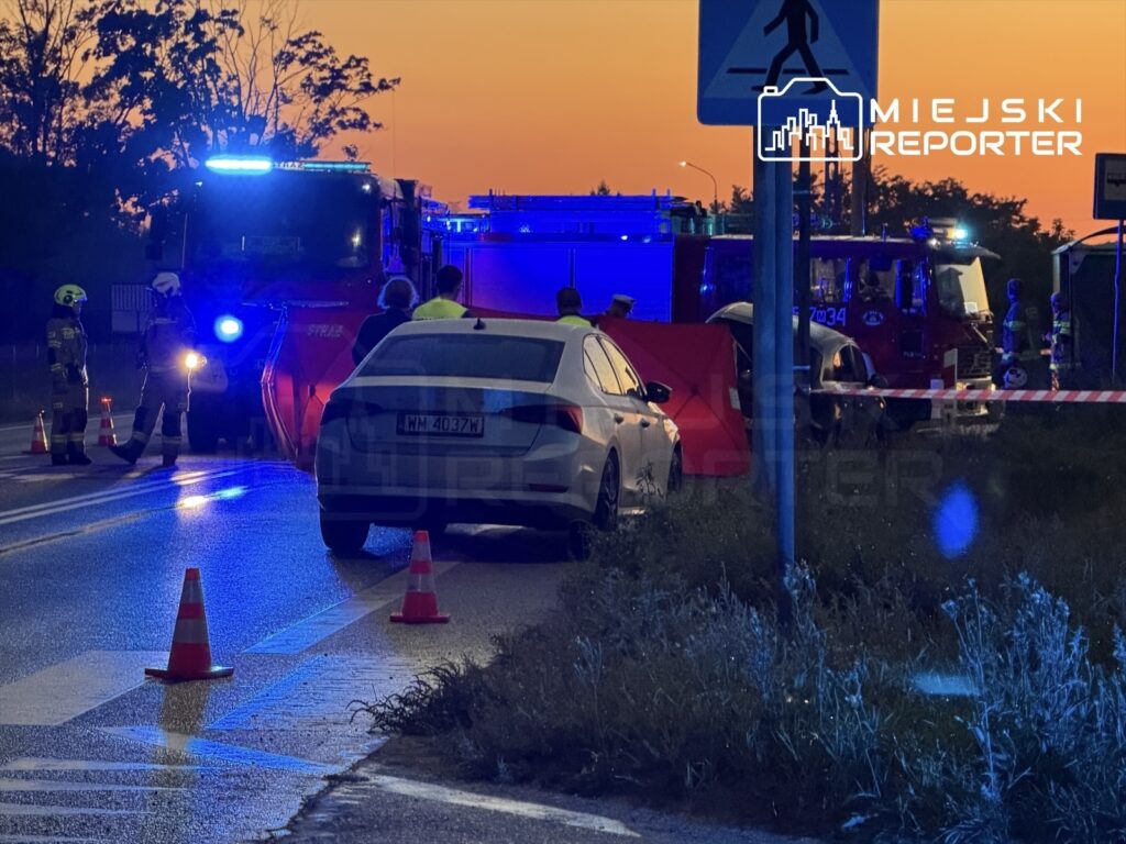 Na drodze, w otoczeniu zachodzącego słońca, strażacy i policjanci pracują przy wypadku, obok czerwonego wozu strażackiego.
