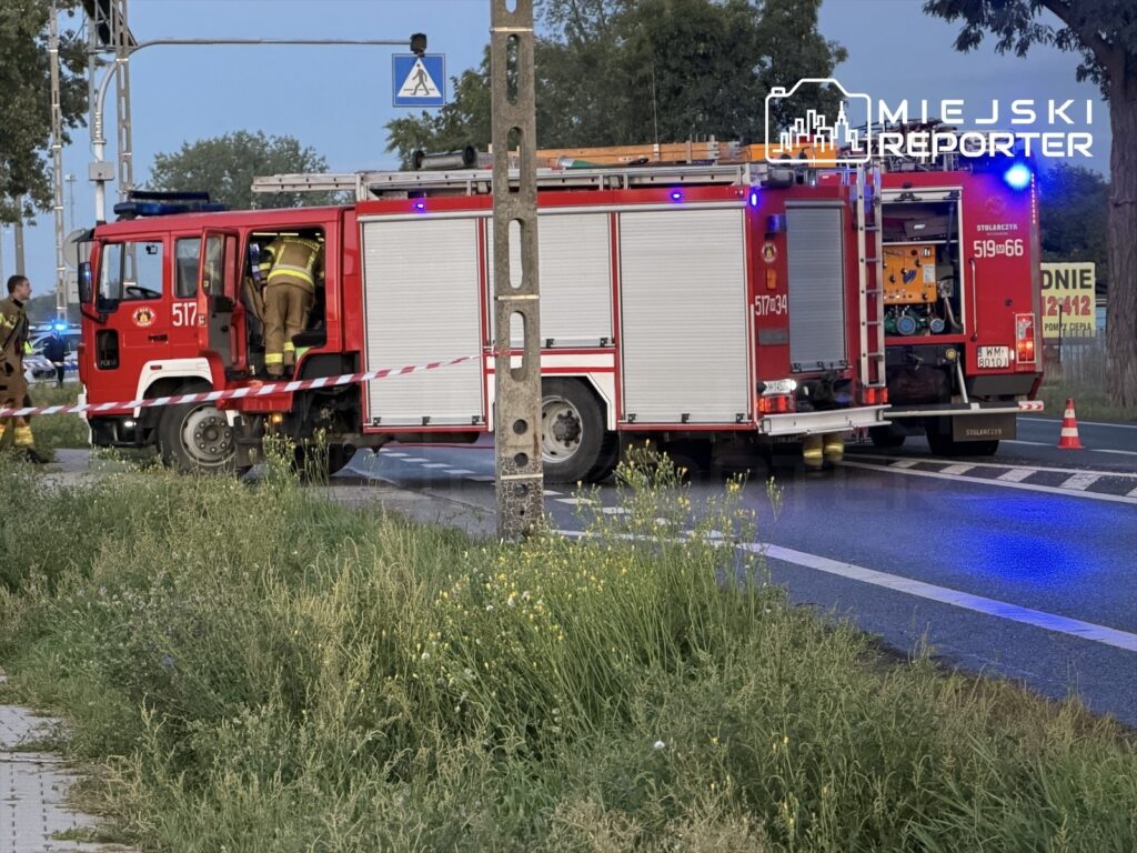 Strażacy w mundurach wsiadają do wozu strażackiego na drodze, obok oznakowania i roślinności przyjezdnej.