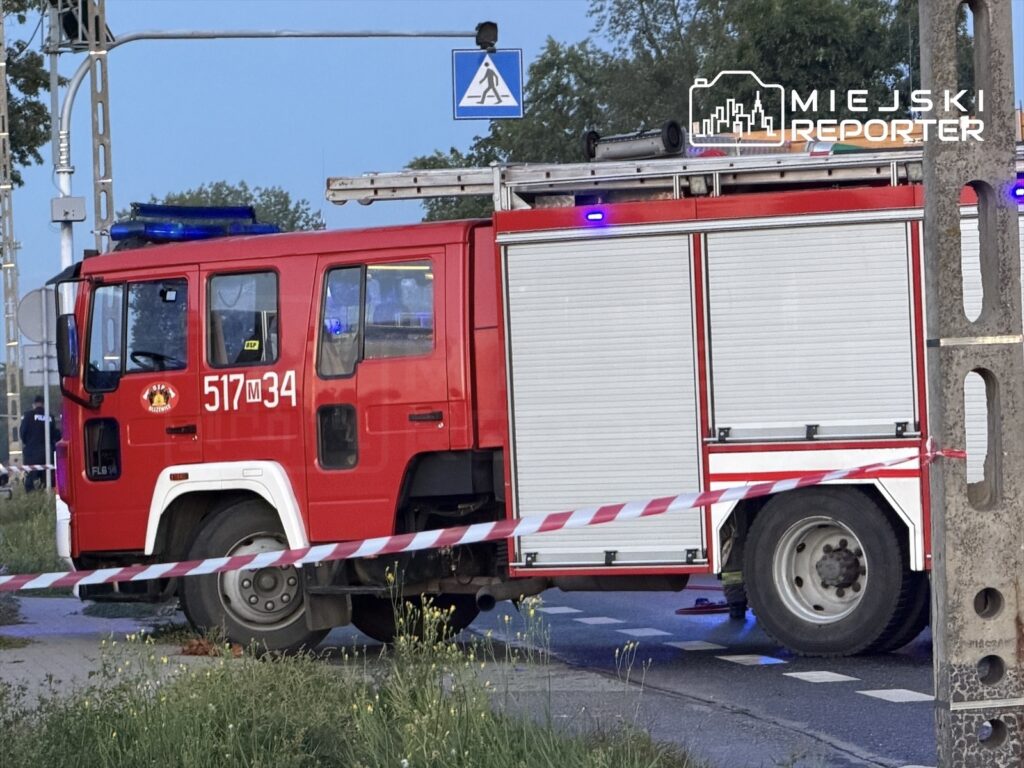 Czerwony wóz strażacki z włączonymi sygnałami świetlnymi stoi przy drodze, otoczony taśmą ostrzegawczą.