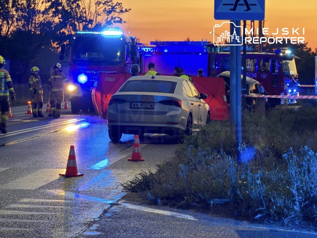 Na drodze stoi samochód osobowy, obok niego pracują strażacy, a w tle widać wozu strażackie z włączonymi sygnałami świetlnymi.