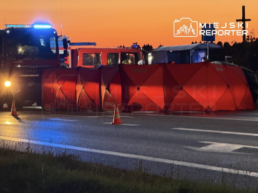 Czerwony namiot zabezpieczający ustawiony na drodze, obok pojazdów straży pożarnej w trakcie akcji ratunkowej.