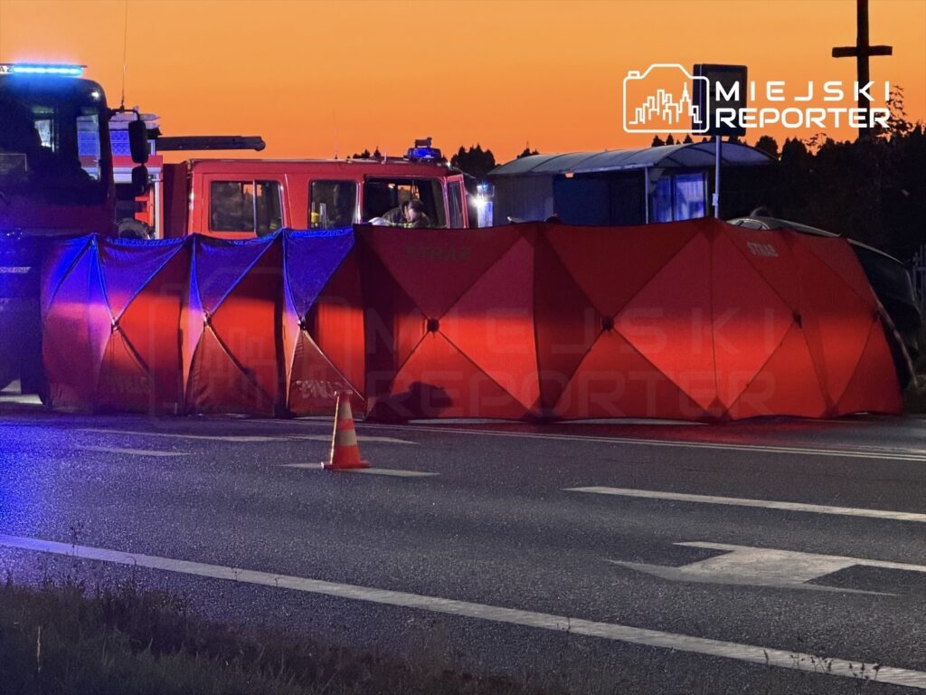 Na drodze ustawione są czerwone namioty, a w tle widoczne są pojazdy straży pożarnej oraz sygnalizacja świetlna.