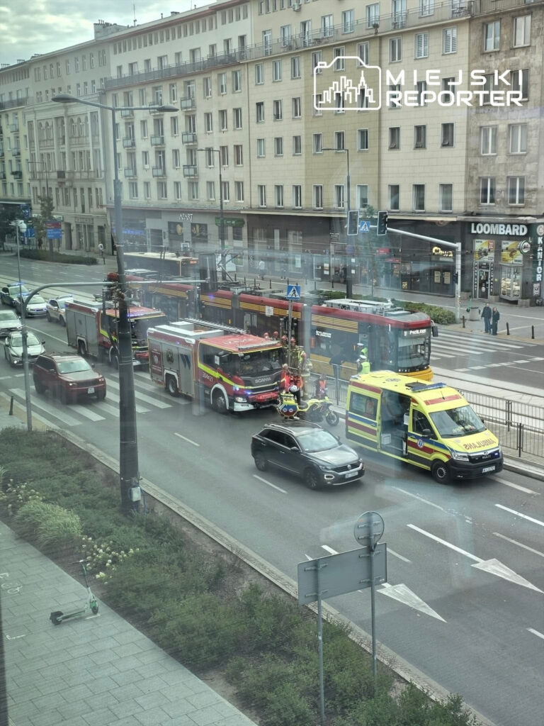 W centrum miasta, w pobliżu przystanku tramwajowego, strażacy i zespół ratunkowy udzielają pomocy na miejscu zdarzenia.