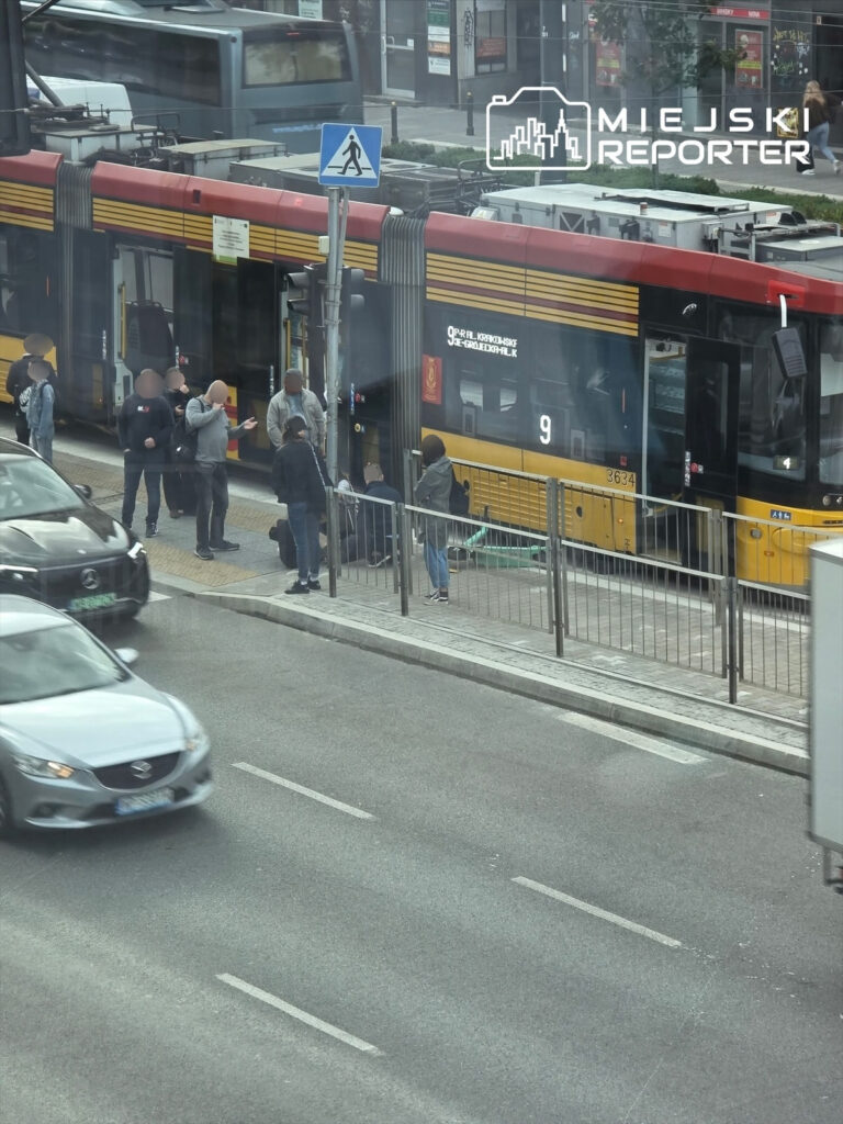 Grupa osób czeka na przystanku tramwajowym obok tramwaju linii 9, w ruchliwej miejskiej ulicy.