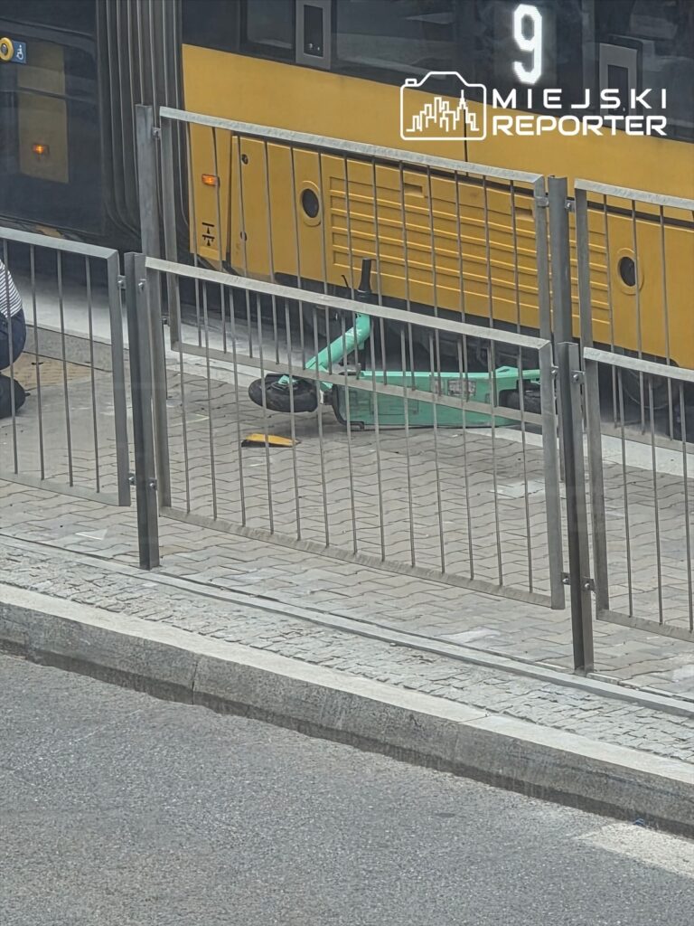 Zielona hulajnoga leży na chodniku obok przystanku tramwajowego, w tle widoczny tramwaj.