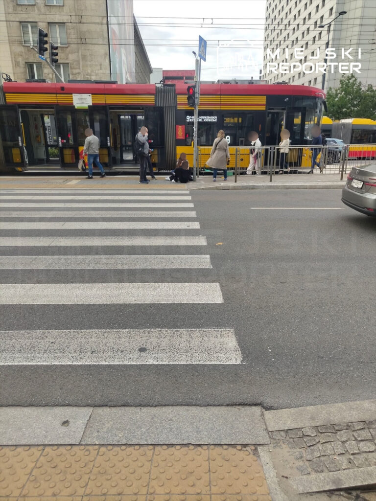 Na przejściu dla pieszych grupa osób obserwuje wypadek, w tle tramwaj zatrzymany na przystanku.