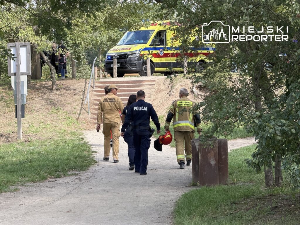 Grupa ratowników i policjantów idzie w stronę schodów w parku, obok pojazdu służb ratunkowych.