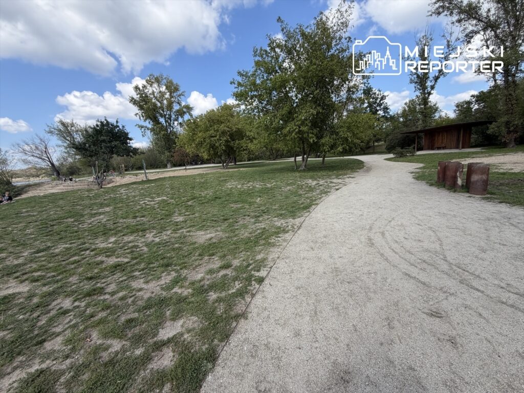 Zielony park z alejkami, drzewami i miejscami do wypoczynku, w tle widoczne huśtawki.