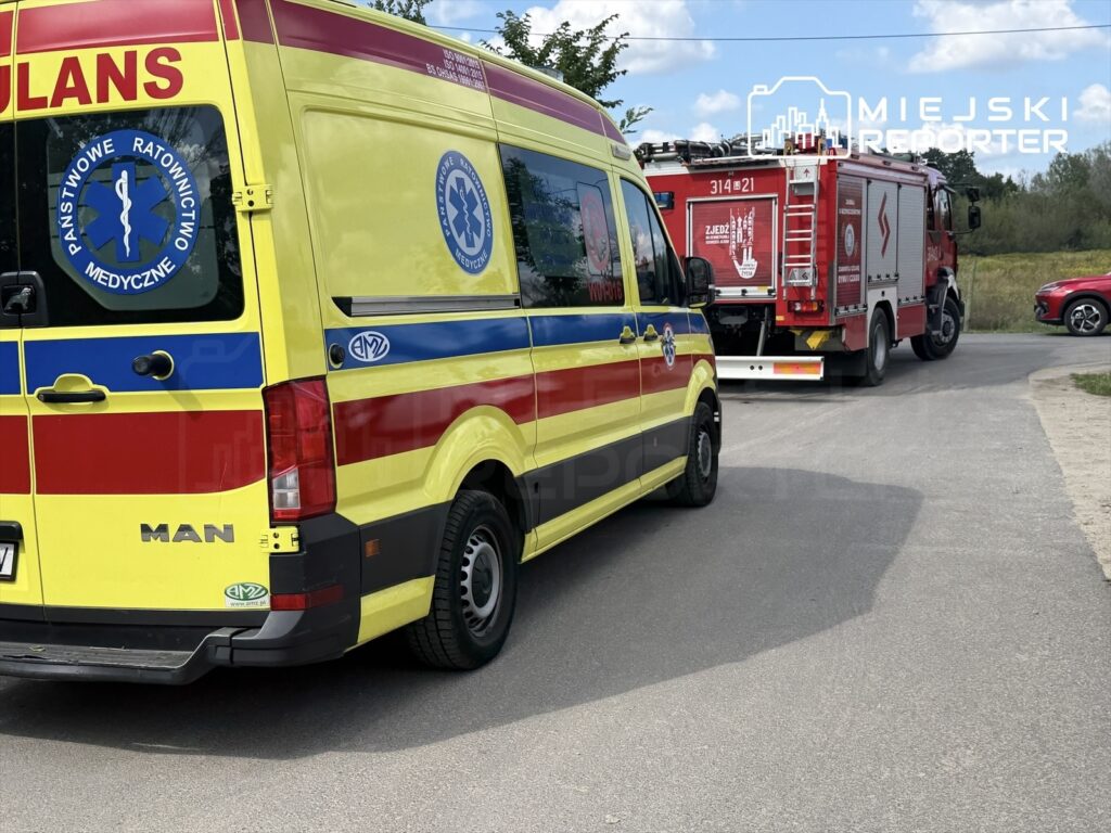 Ambulans w kolorze żółto-czerwonym stoi na drodze, obok niego widać wóz strażacki. W tle rosną drzewa.