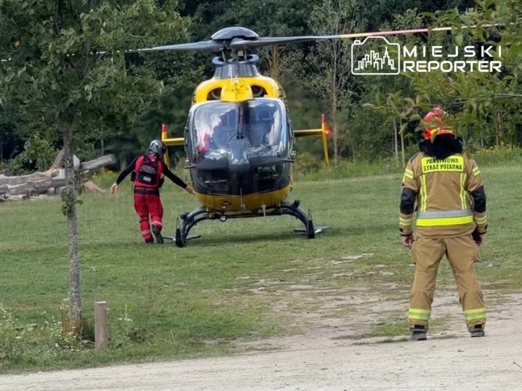 Ratownik w czerwonym stroju zbliża się do lądowiska helikoptera, obok stoi strażak w mundurze.