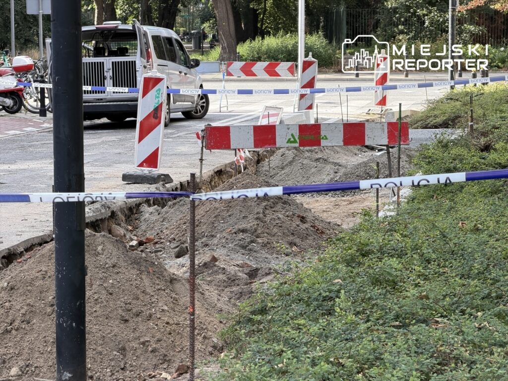 Wyznaczony obszar robót drogowych z wykopem, oznaczony barierkami i taśmą ostrzegawczą. W tle widoczny samochód służbowy.