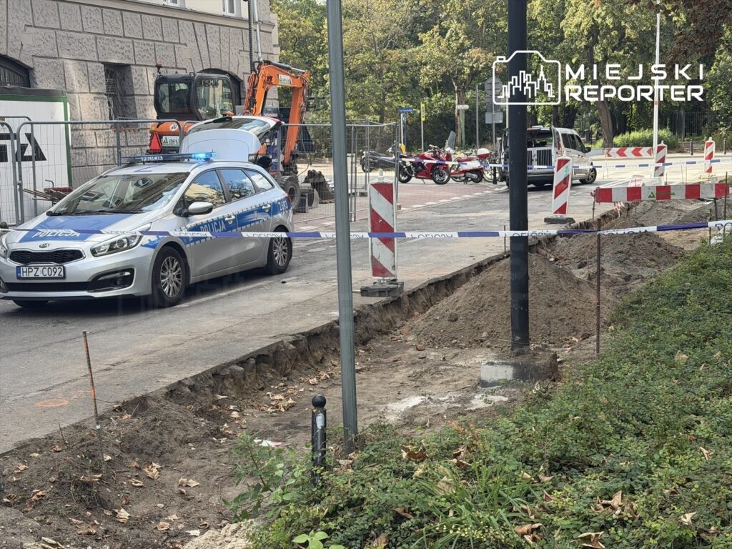 Policyjny radiowóz stoi obok wykopu na ulicy, zabezpieczonego taśmą. W tle widać sprzęt budowlany i znaki drogowe.