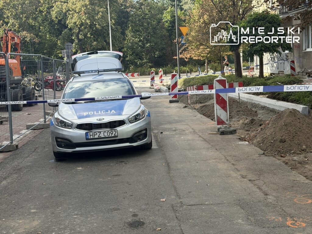 Policyjny radiowóz stoi na ulicy, otoczony barierkami budowlanymi, w tle prace drogowe.