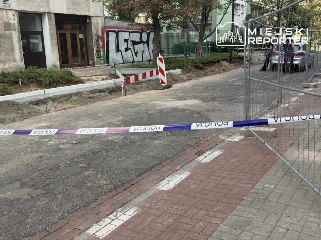 Robotnicy prowadzą prace budowlane przy chodniku, ogrodzeni plac robót zabezpieczony taśmą policyjną.