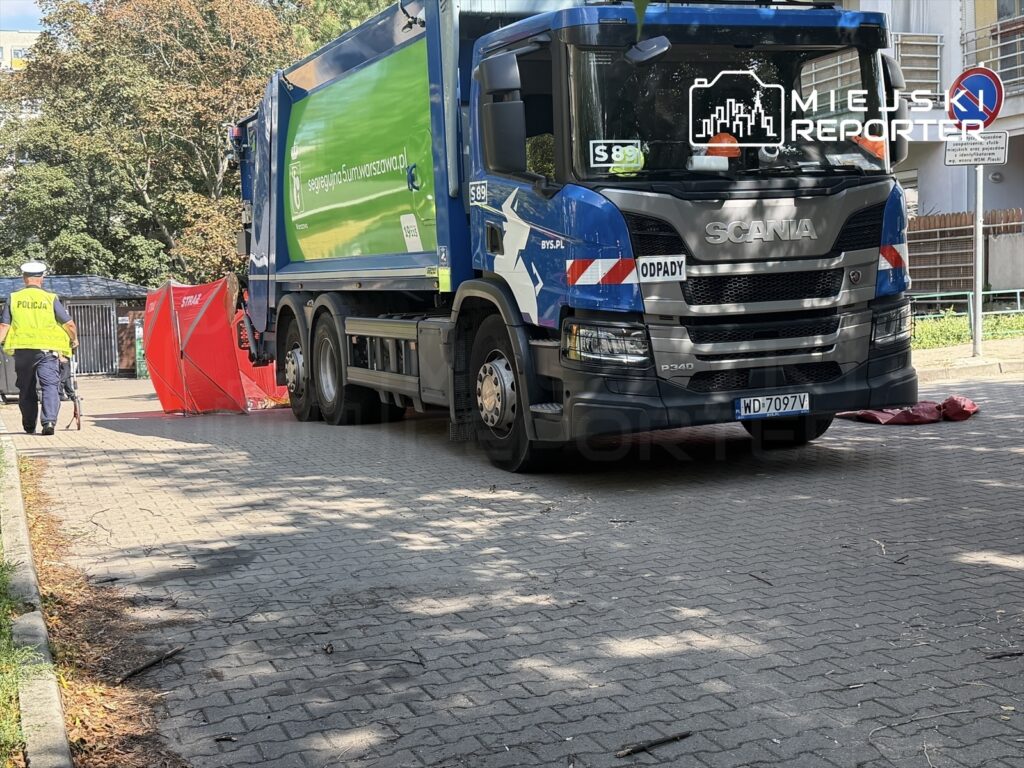 Policjant w kamizelce odblaskowej przechodzi obok śmieciarki, a w tle widoczny jest czerwony namiot zabezpieczający.