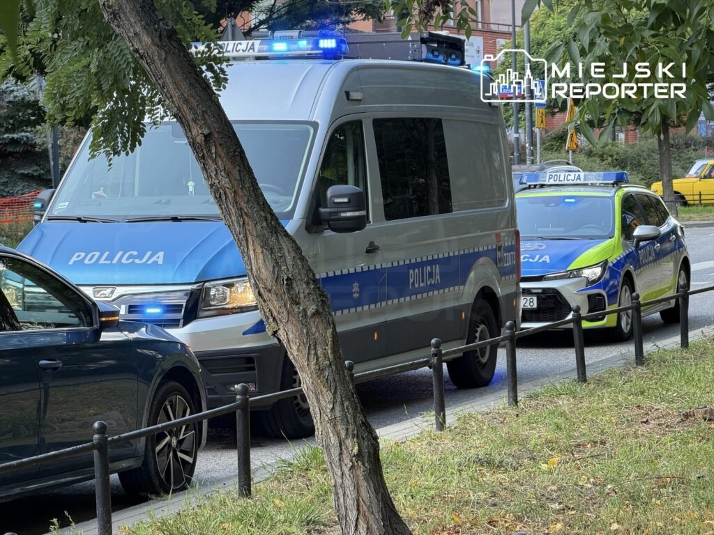 Policyjny radiowóz i furgonetka zaparkowane na ulicy, z włączonymi sygnałami świetlnymi.