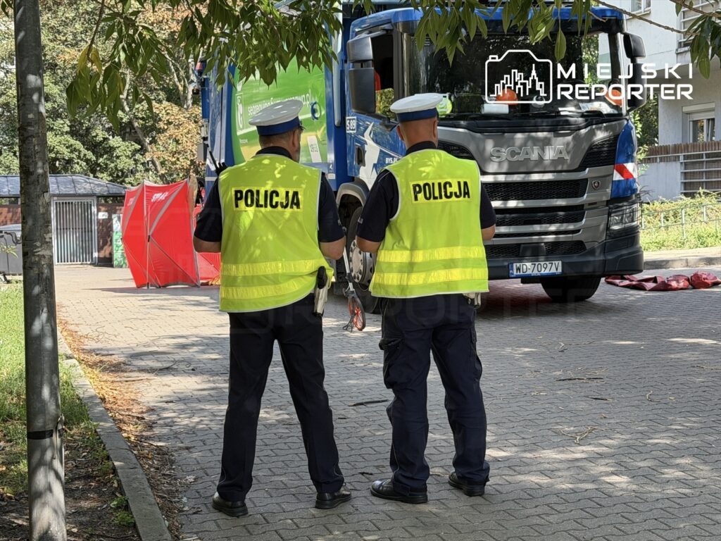 Dwaj policjanci w kamizelkach odblaskowych obserwują akcję w pobliżu dużego niebieskiego ciężarowego pojazdu.