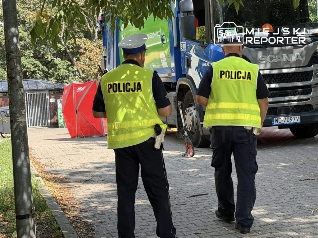 Dwóch policjantów w odblaskowych kamizelkach obserwuje teren w pobliżu niebieskiego ciężarowego pojazdu.