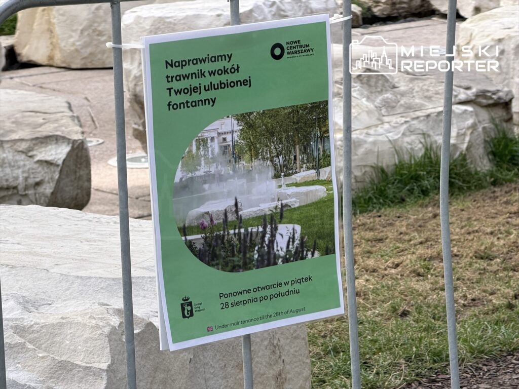 Plakat informacyjny na ogrodzeniu z napisem "Naprawiamy trawnik wokół Twojej ulubionej fontanny" obok kamieni.